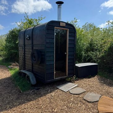 A small black wooden cabin trailer with a chimney in a natural setting.