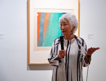 Elderly woman speaking in front of abstract art at an exhibit.