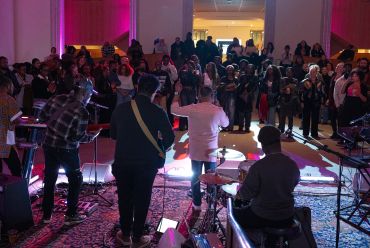 A band performs live indoors for a standing audience in colorful lighting.