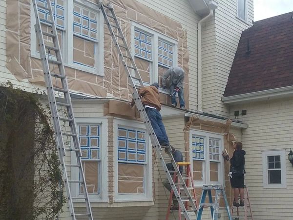 Workers preparing a house exterior for painting with ladders and masking.