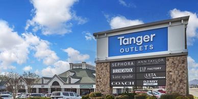 Tanger Outlets shopping center with various brand signs under a blue sky.
