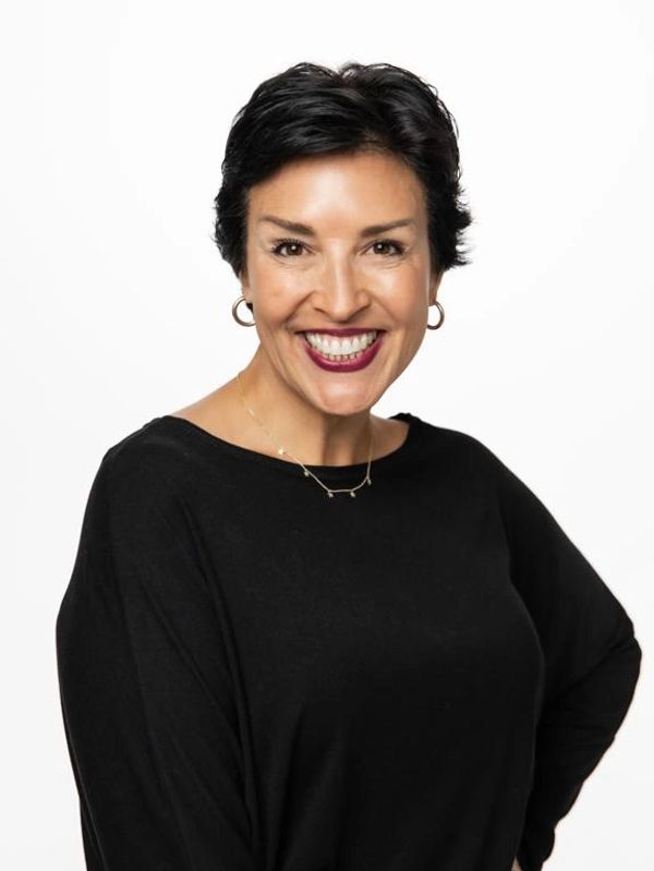 Confident woman with short black hair smiling warmly, wearing a black top and gold hoop earrings.