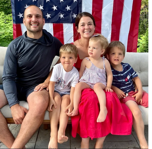 Smiling family of five sitting in front of an American flag outdoors.