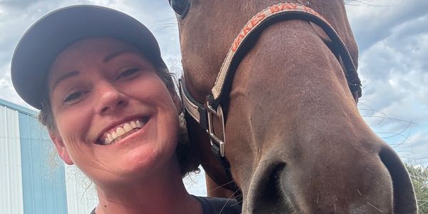 Smiling woman takes a close selfie with a brown horse outdoors.