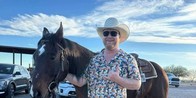 A man in a cowboy hat posing with a horse under a partly cloudy sky.