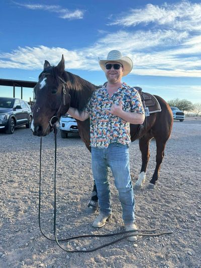 Man in a cowboy hat poses with a saddled horse under a blue sky.