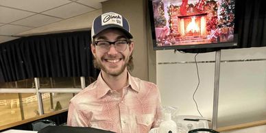Smiling man wearing a cap and glasses indoors with a festive fireplace on TV.