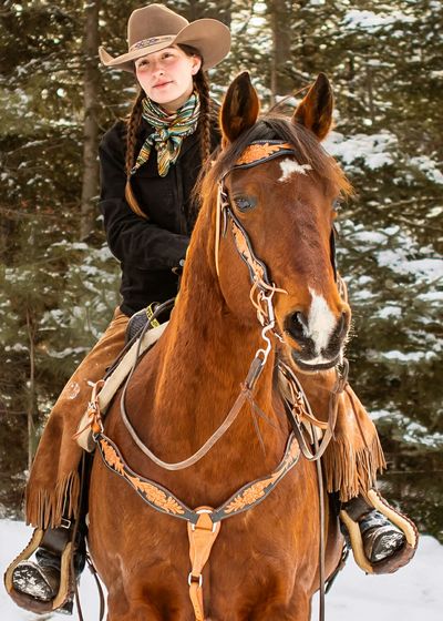 Young woman dressed as a cowboy riding a brown horse in a snowy forest.