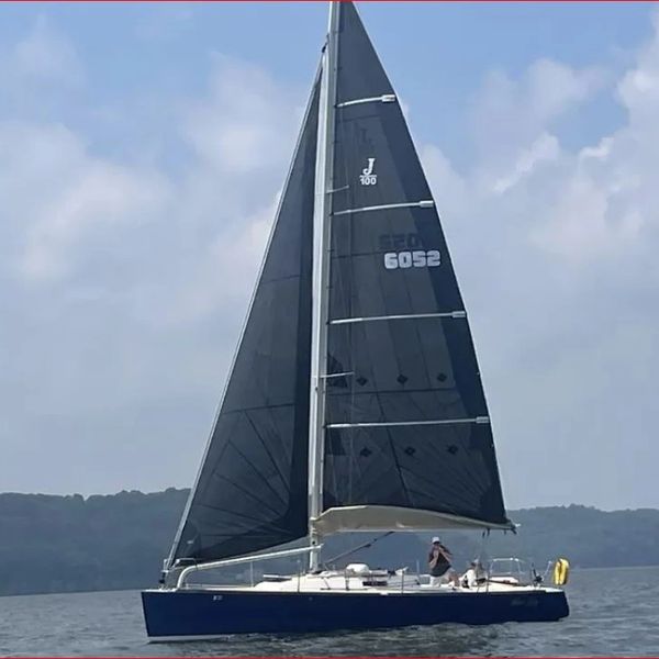 A sleek sailboat with black sails glides on calm water under a cloudy sky.