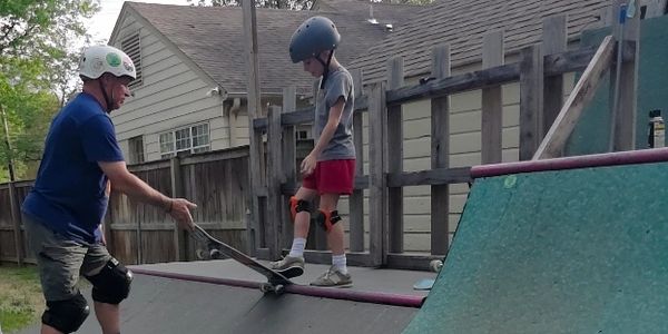 Adult teaching child to skateboard on a backyard ramp.