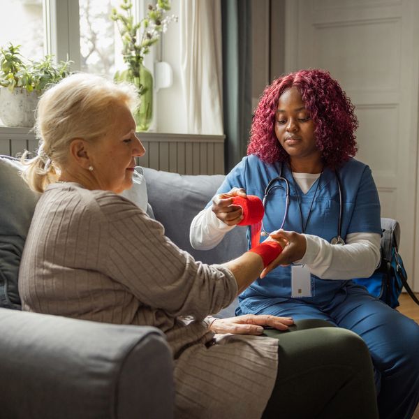 Healthcare worker wrapping an elderly woman's hand with a red bandage at home.