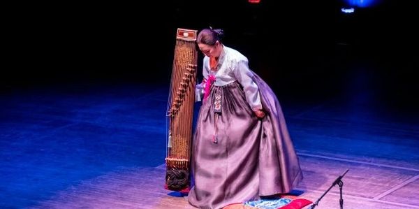 A woman in traditional Korean hanbok bowing on stage next to a gayageum.