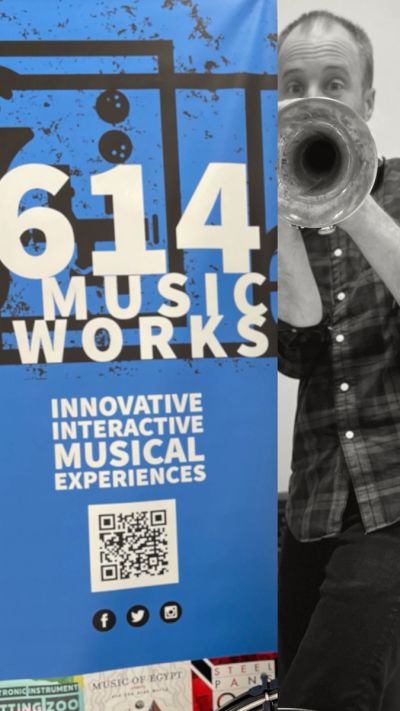 Man playing trumpet next to 614 Music Works banner.