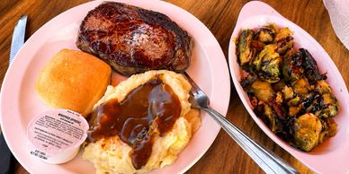 A hearty meal with steak, mashed potatoes with gravy, roll, and a side of roasted Brussels sprouts.