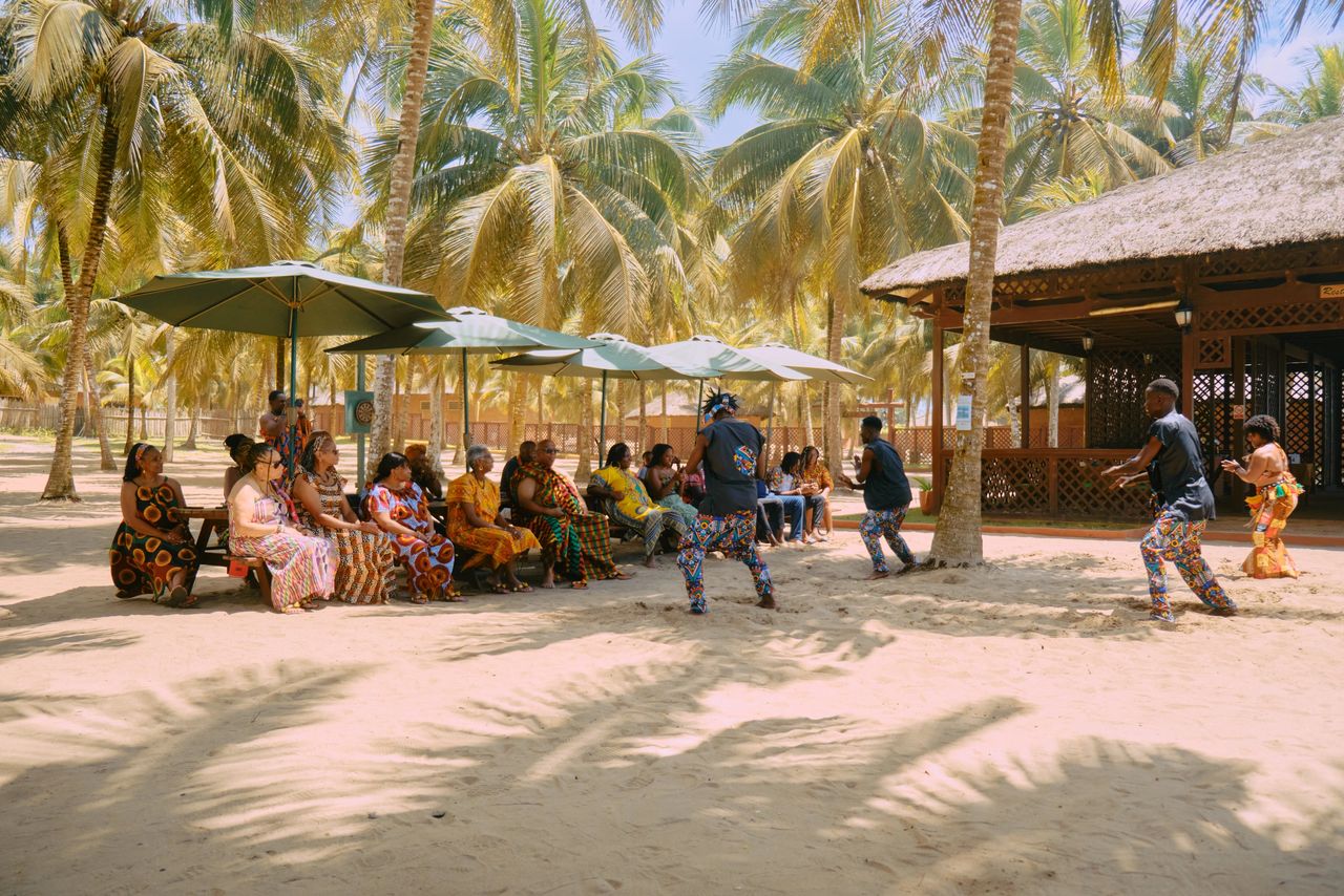 The image portrays a tropical beach setting with tall coconut palm trees swaying in the background, their fronds providing dappled shade on the sandy ground below. Beneath the trees, a group of individuals, primarily women, is seated on a long bench under a large umbrella. The women are dressed in vibrant, colorful traditional outfits, featuring patterns reminiscent of African prints. Their attire, adorned with an array of hues including red, yellow, blue, and orange, stands out prominently against the natural backdrop. In the foreground, three individuals are actively engaged in what seems to be a cultural or traditional performance. One person, donned in a blue outfit with bold patterns, seems to be dancing or moving towards the seated group, while the other two, one in a similar patterned outfit and the other in a bright orange dress, are moving in the opposite direction. Their dynamic poses suggest motion and activity. Adjacent to the gathering is a thatched-roof structure, possibly a hut or shelter, which further enhances the tropical ambiance of the scene. The atmosphere exudes a sense of community, tradition, and celebration, capturing a candid moment of cultural expression against the serene backdrop of a coastal environment.