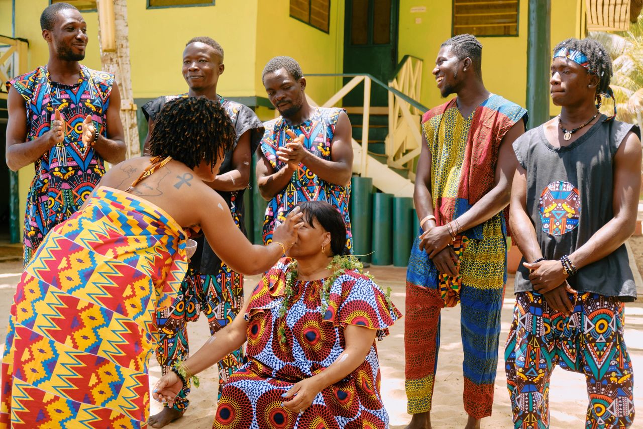 The image captures a vibrant scene from a naming ceremony in Ghana. In the foreground, a woman dressed in a brightly-colored, geometric-patterned outfit leans over another seated woman, who is adorned in similar traditional attire, complemented by green beaded necklaces and an elegant head wrap. The seated woman has a gentle, appreciative smile, as the other woman appears to perform a ritual or offer a blessing. Surrounding them, men dressed in equally vivid and patterned outfits are seen clapping and partaking in the ceremony, with some wearing painted or tattooed symbols on their arms and bodies. The overall ambiance is celebratory and deeply rooted in cultural tradition.