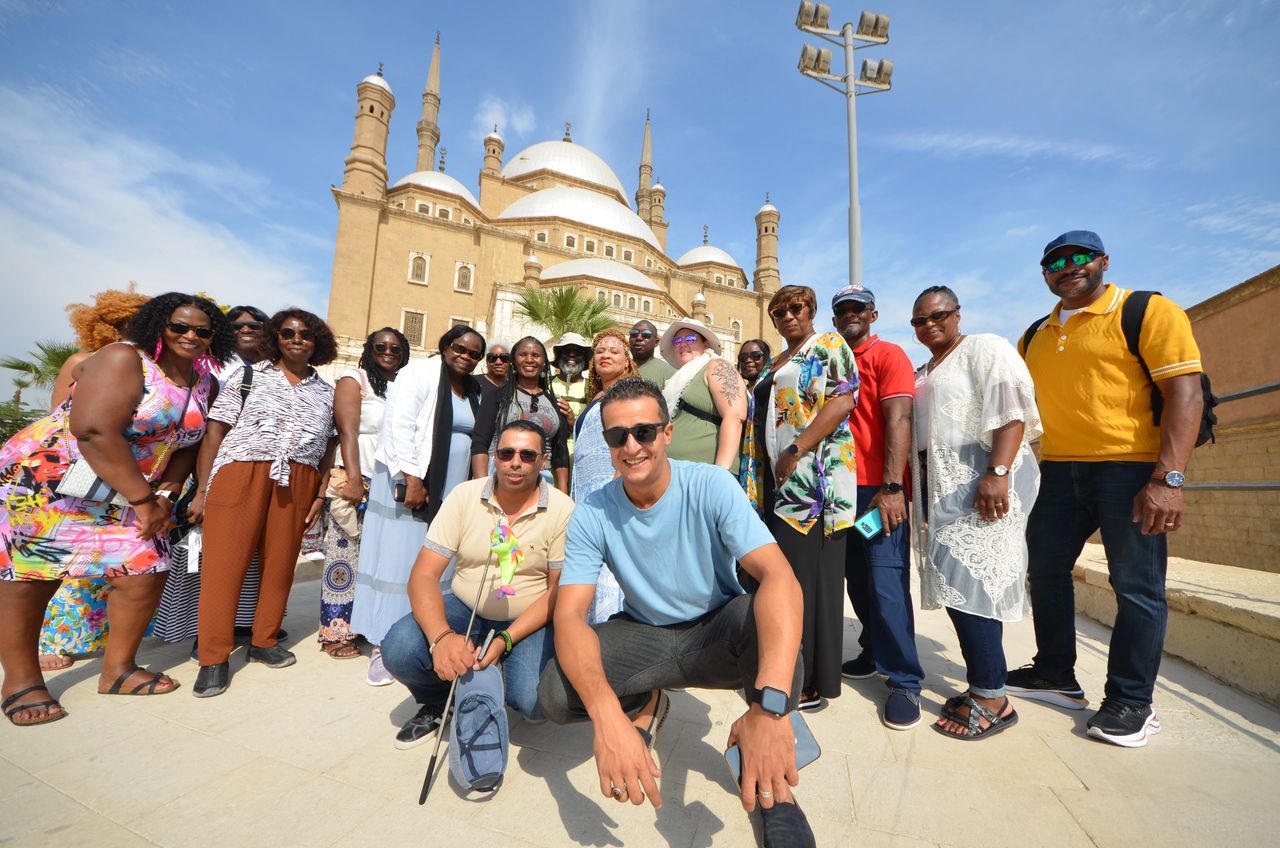 The image showcases a lively group of tourists in Egypt, gathered in front of a majestic mosque with towering minarets and grand domes. The architecture of the mosque is emblematic of traditional Egyptian religious structures. The tourists exhibit a mix of attire, from vibrant summer dresses to casual wear, suggesting a warm climate. Their cheerful expressions reflect the joy of exploration. In the foreground, two men, one holding a selfie stick and the other kneeling with a wide grin, seem particularly engaged in capturing memories.