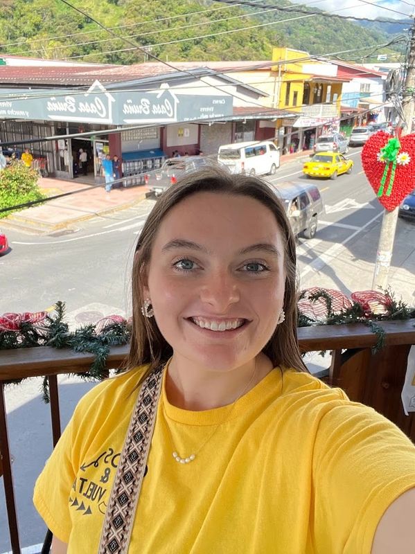 Smiling woman in a yellow shirt taking a selfie outdoors on a sunny day.