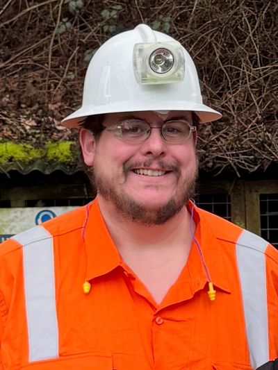 Man in safety gear with an orange vest and white helmet smiling outdoors.