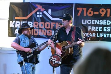 Two musicians perform on stage with guitars in front of a roofing company banner.