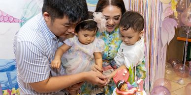 A family celebrates a toddler's first birthday with a decorated cake.