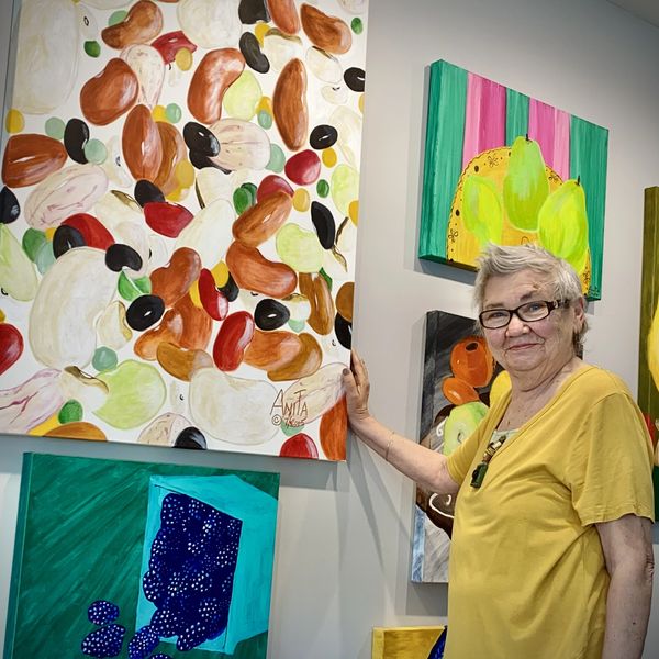 An elderly woman stands next to colorful abstract paintings in a gallery.