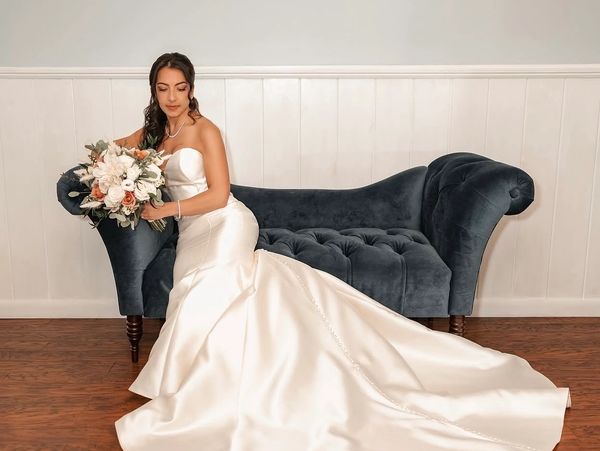 Bride in white gown sitting on dark velvet sofa.