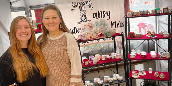 Two smiling women at a cozy candle and melts market booth.