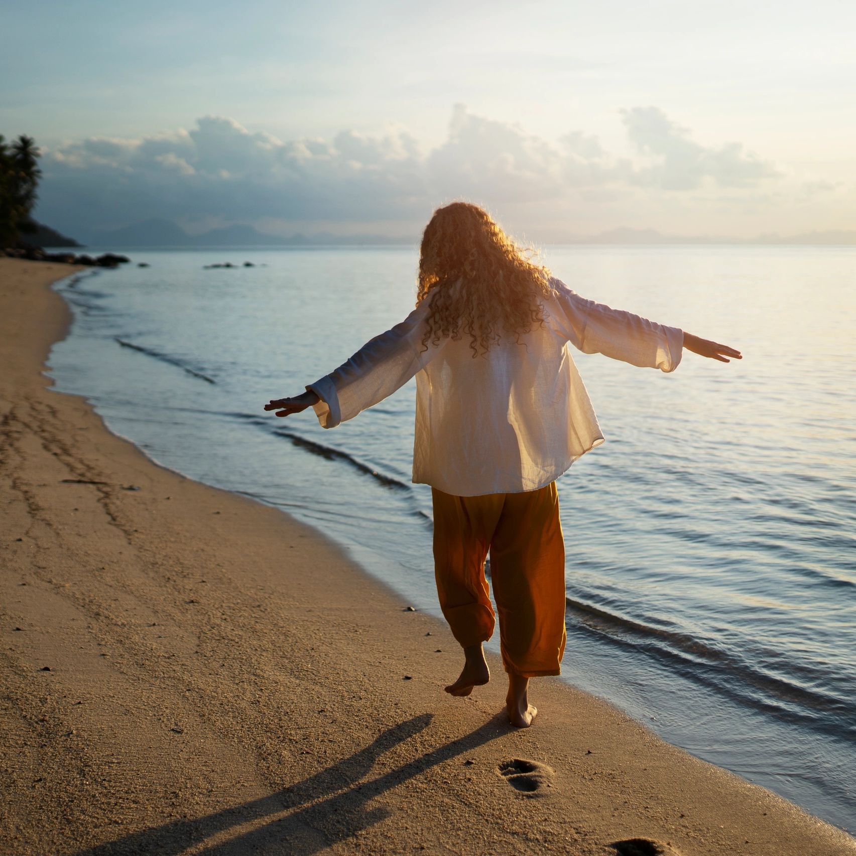 Person with curly hair walking barefoot on a beach at sunrise.