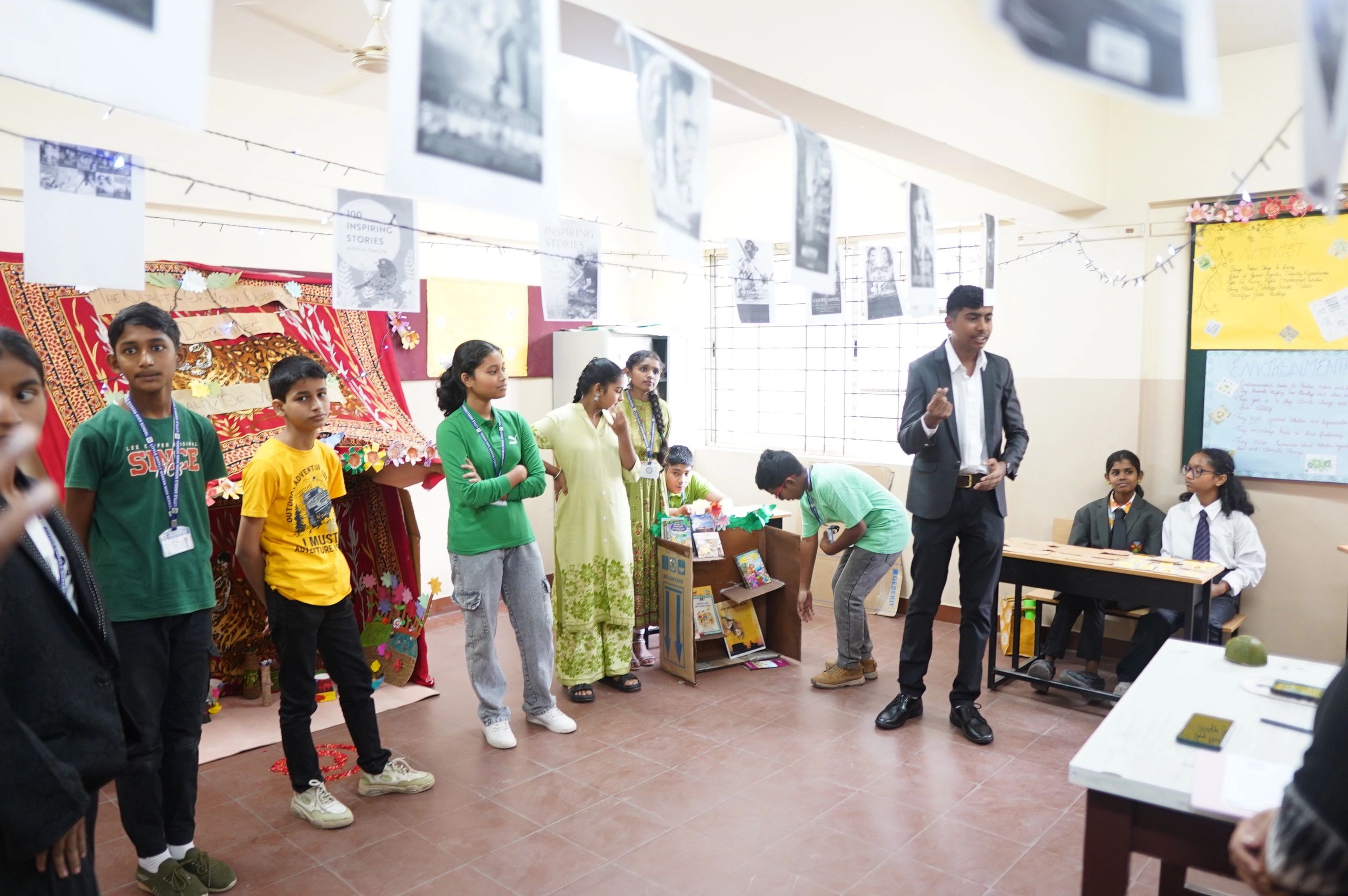 Students presenting and interacting in a classroom decorated with hanging photos and posters.