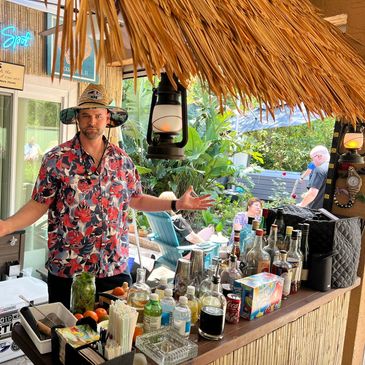 Man in tropical shirt at a tiki bar with various drinks and decorations.