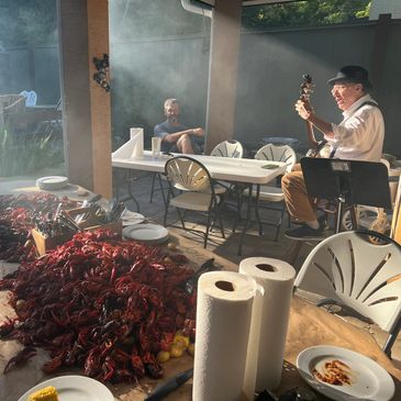 Man playing guitar at a crawfish boil with piles of crawfish and people relaxing.