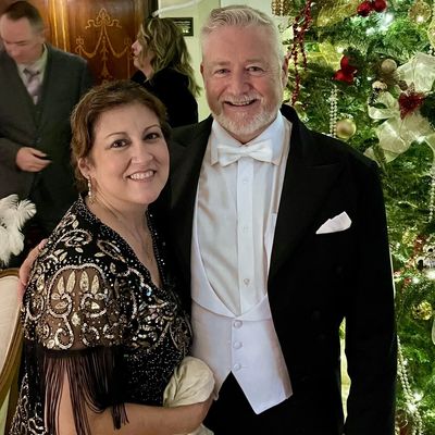A smiling couple dressed elegantly in front of a decorated Christmas tree.