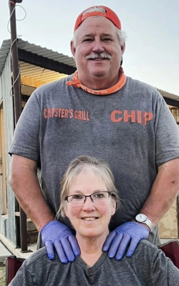 A smiling man stands behind a seated woman, resting his gloved hands on her shoulders.