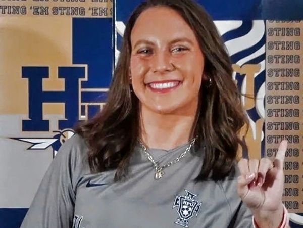 A smiling female athlete in a gray sports jersey making a hand gesture.