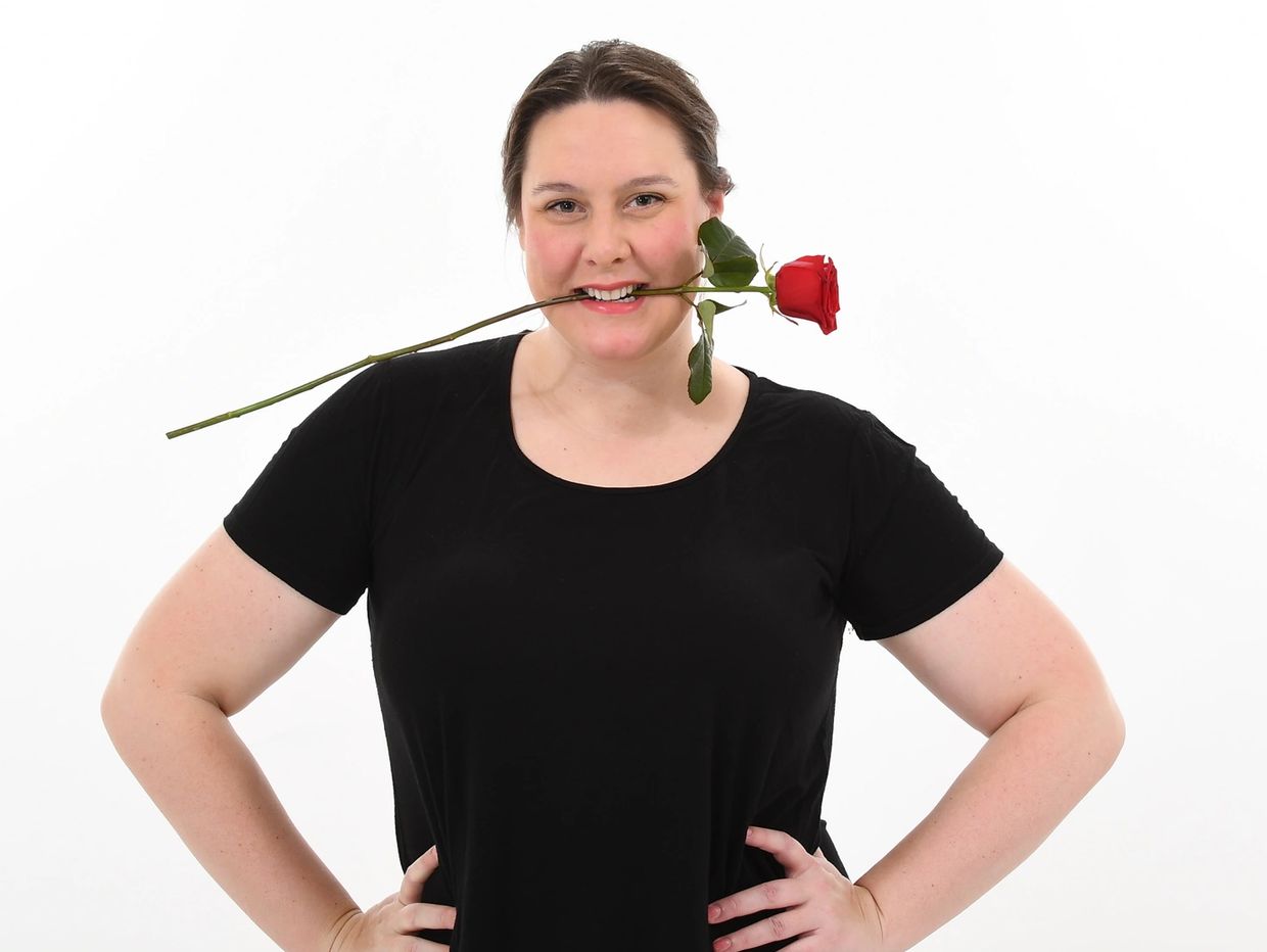 Woman in black shirt holds a red rose in her mouth, smiling.