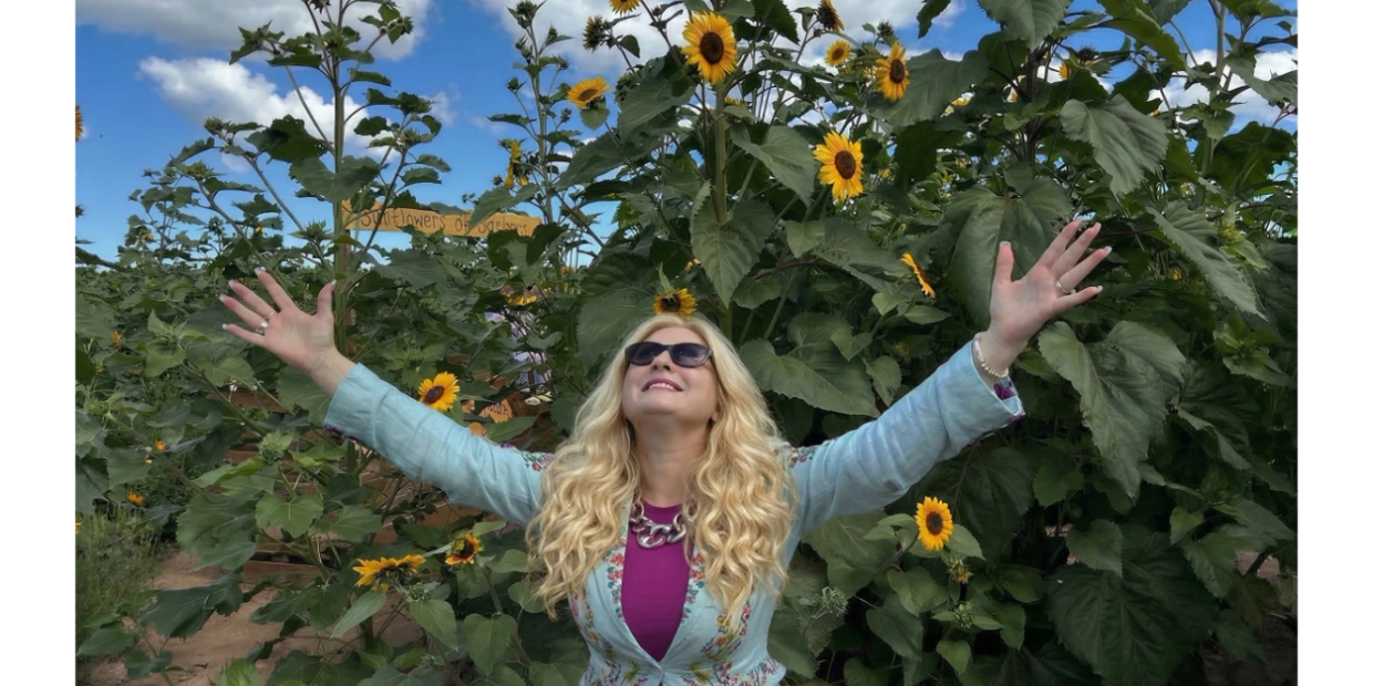 Woman joyfully spreading arms in front of tall sunflowers under blue sky.