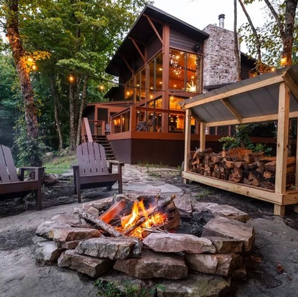 Cozy fire pit with wood storage and a glass-walled cabin in the woods.