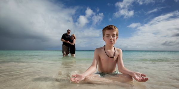 A boy meditates in shallow ocean water with a couple embracing in the background.