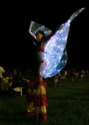 Performer on stilts with illuminated butterfly wings at night.