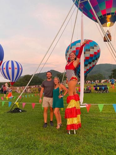 Two people standing with a performer on stilts at a hot air balloon event.