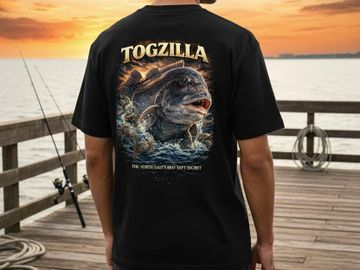 Person wearing a black Togzilla graphic tee on a pier at sunset.