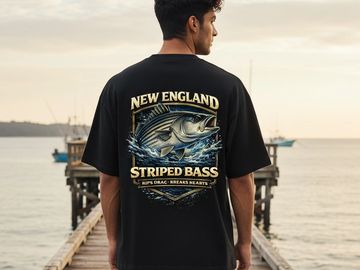 Man wearing a black T-shirt with a New England striped bass design on a dock by the water.