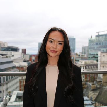 Woman with long dark hair standing on a rooftop with city buildings behind.