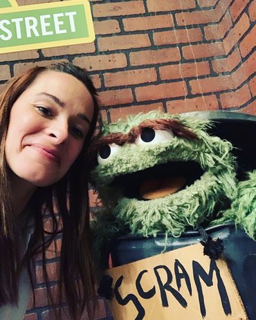 Woman smiling next to Oscar the Grouch puppet from Sesame Street.