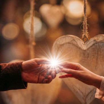 Two hands touching with a glowing light between them and heart decorations in background.