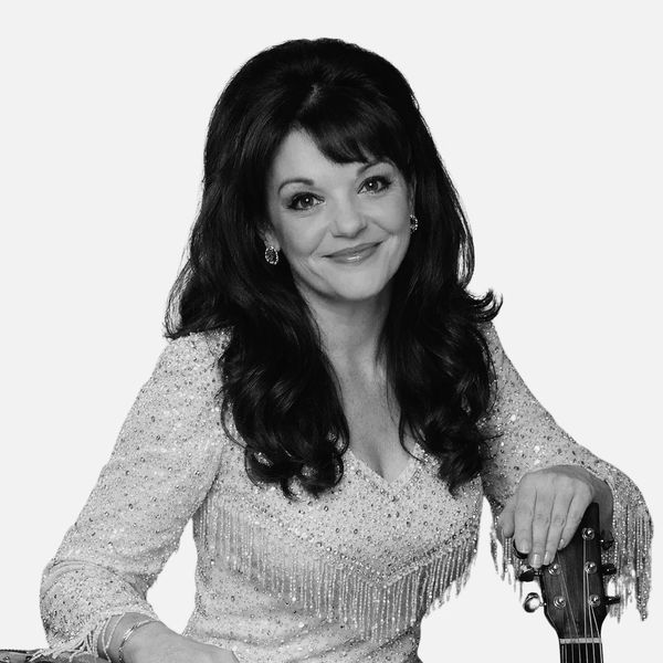 Smiling woman with long dark hair in a sparkly dress holding a guitar.