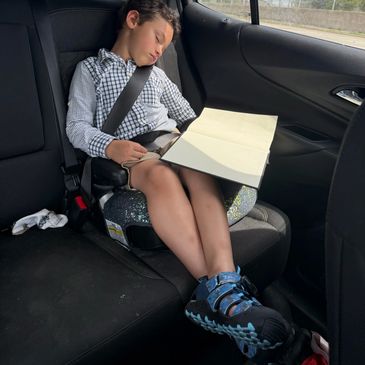 A young boy sleeping in a car seat with an open book on his lap.