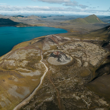 Aerial view of a volcanic landscape with a lake and rugged terrain under a partly cloudy sky.