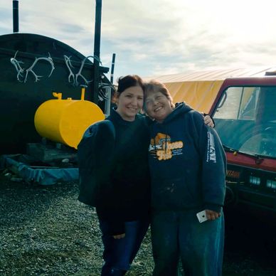 Two women smiling and hugging near a rustic building and a red vehicle outdoors.